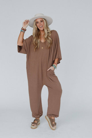 A smiling woman with long blonde hair wears a wide-brimmed white hat, the Lovely As Ever Harem Jumpsuit from Three Bird Nest in an oversized boho brown style, and tan sandals. She accessorizes with bracelets on both wrists and holds onto her hat with her right hand. The background is plain light gray.