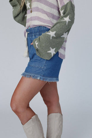 A person is wearing a green sweater with white stars over a striped pink and white shirt, paired with the Meet The Moment Drawstring Shorts in Dark Wash by Three Bird Nest. They complete the outfit with cream-colored knee-high boots and are posing against a plain light gray background.
