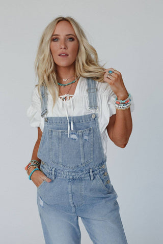 A woman with long blonde hair stands against a plain background, wearing a white blouse and the Cali Dream Denim Overalls in Light Wash by Three Bird Nest, crafted from comfortable fabric. She accessorizes with multiple necklaces, colorful bracelets, rings, and has her left hand in her pocket, exuding a boho-chic vibe.