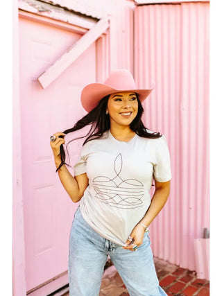 A woman wearing a pink cowboy hat, the Ellery Boot Stitch Tee from Western Edge Boutique, and light blue jeans stands in front of a pastel pink building. She is smiling and holding a lock of her long dark hair, which flows down over her shoulders.