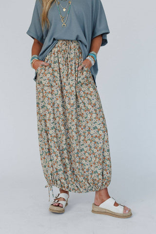A person wearing a loose, blue-gray top tucked into a Three Bird Nest Solstice Song Cinched Skirt in Khaki, which features small flowers in various colors. The high-rise, floral maxi skirt has pockets where their hands are tucked. They are also wearing white sandals and several bracelets.