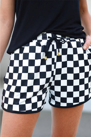 A person is wearing Roxy Women's Checkered Drawstring Elastic Waist Shorts by Aapparella, featuring a comfortable black and white checkered design with a black drawstring accented by gold tips. They also sport a black top and have one hand casually placed in a pocket. The blurred background lends an indistinct charm to their effortless summer style.