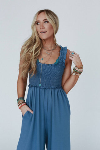 A woman with long, wavy blonde hair is wearing the Nothing Compares Smocked Ruffle Jumpsuit - Denim Blue by Three Bird Nest, made of lightweight stretch fabric. It features a ruffled neckline and a shirred bodice. She accessorizes with layered necklaces and multiple bracelets, casually posing with one hand in her pocket against a plain background.
