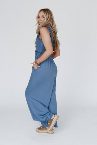 A woman with long, wavy blonde hair stands in a side pose, wearing the "Nothing Compares Smocked Ruffle Jumpsuit" in denim blue from Three Bird Nest. The jumpsuit features ruffled shoulders and a shirred bodice made from lightweight stretch fabric. She has multiple bracelets on her right wrist and is wearing tan platform sandals. The background is plain and white.