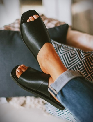 Close-up of a person's feet wearing Stormi Black Slide Sandals from Kay Kay Fashion. Their ankles are crossed and resting on a cushioned surface, showcasing women's flat shoes that are both comfortable and true to size. The person is wearing blue rolled-up jeans, with patterned cushions and soft furnishings in the background.