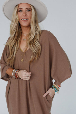 A woman with long blonde hair wearing a wide-brimmed hat and the "Lovely As Ever Harem Jumpsuit" by Three Bird Nest, stands with one hand in her pocket. She accessorizes with multiple bracelets and layered necklaces, smiling slightly against a plain gray background.