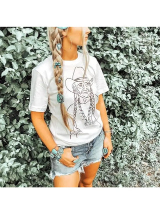 A woman stands outdoors wearing the Willie "Original Hippie" Tee from female-owned brand Hank & Scoot. The white t-shirt features a sketch of a person in a cowboy hat and braid, paired with denim shorts and turquoise jewelry. Her long braided hair, adorned with turquoise accessories, embodies the rustic charm amidst Texas greenery. Available in unisex sizing, this shirt perfectly captures her bohemian style.