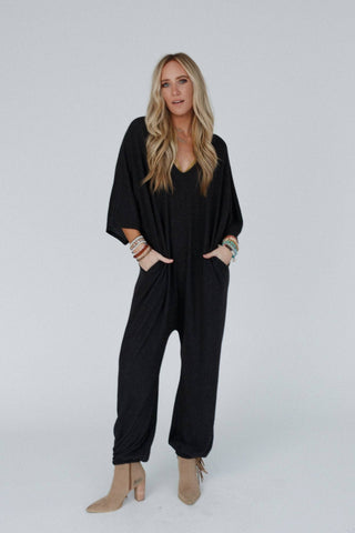 A woman with long blonde hair stands against a plain background, wearing the comfy Lovely As Ever Harem Jumpsuit in charcoal by Three Bird Nest, with her hands in the pockets. She accessorizes with several boho bracelets on both wrists and tan ankle boots. She gazes at the camera with a neutral expression.