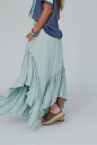 A woman is adorned in the elegant Logan Forever Beauty Maxi Skirt in Pale Sage from Three Bird Nest, showcasing its flowing fabric and ruffle details. She complements her look with taupe sandals and a comfortable high-rise elastic waistband that ensures a perfect fit. The lightweight stretch knit fabric allows for easy movement, while she pairs the skirt with a coordinating blue top and multiple stacked bracelets. Her hands rest casually inside the skirt's convenient pockets.