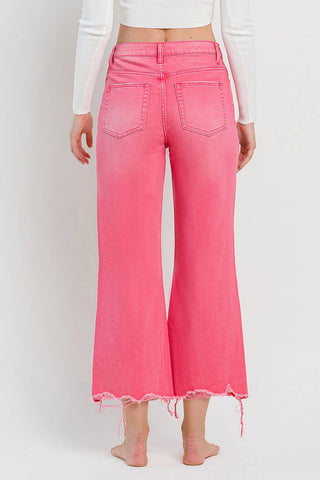 A person wearing VERVET by FLYING MONKEY's Rosie 90's Super High Rise Crop Flare Hot Pink Jeans and a white long-sleeve top stands with their back to the camera against a plain white background. The high-waisted jeans, featuring back pockets and a vintage 90's flare, have a frayed hem. The person is barefoot.