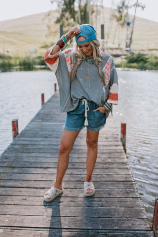 A person stands on a wooden dock by a lake, wearing a blue cap backward, a grey long-sleeve top with colorful patterns, Meet The Moment Drawstring Shorts - Dark Wash from Three Bird Nest, and sliders. They smile while holding the edge of their cap with one hand. Trees and hills are visible in the background.