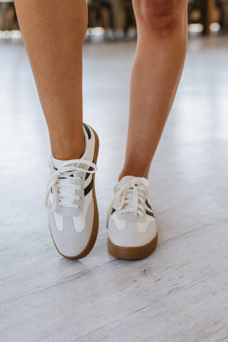 Close-up of a person standing feet together, slightly inward on a light wood floor, wearing Liam & Company Ness Striped Lace Up Flat Sneakers featuring gum soles and neatly tied black stripes.