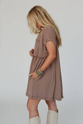 A person with long blonde hair, wearing the Meadow Subtle Escape Textured Dress by Three Bird Nest that exudes boho western vibes, stands sideways. They have one hand in a pocket and wear several colorful beaded bracelets. Complementing the ensemble are light-colored boots, set against a plain light gray background.