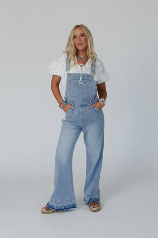 A woman with long blonde hair wears a white blouse under the Cali Dream Denim Overalls - Light Wash by Three Bird Nest, made of comfortable fabric. She has her hands in the pockets of the overalls. Adorned with various boho bracelets and wearing sandals, she stands against a plain white background.