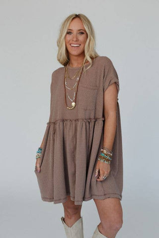 A woman with wavy blonde hair wears a loose-fitting, short-sleeved Meadow Subtle Escape Textured Dress from Three Bird Nest that exudes boho western vibes. She accessorizes with layered necklaces and multiple bracelets, standing and smiling against a plain background.