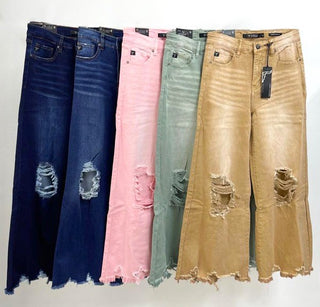 A display of five pairs of Calico Camel Distressed Crop Flare jeans by Lucky & Blessed in various colors: dark blue, light blue, pink, mint green, and beige. Each pair features high-rise tummy control with frayed hems and prominent rips on the knees and thighs, hanging side by side.