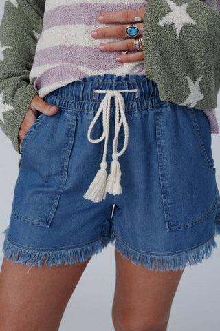 A person is wearing the "Meet The Moment Drawstring Shorts - Dark Wash" by Three Bird Nest, which feature blue denim with frayed edges and a white drawstring waistband. Their hand, adorned with multiple rings, rests on their hip. They are also sporting a cozy long-sleeve sweater that has green sleeves and white star patterns.