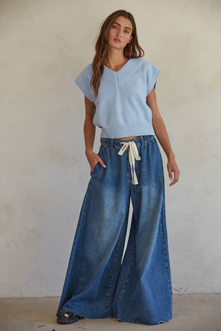 A woman with long brown hair is wearing a light blue sleeveless sweater and Brynlee Denim Waist Band Wide Leg Pants in Dark Denim by By Together. She is standing against a neutral background with her hands in her pockets and looking slightly to the side.