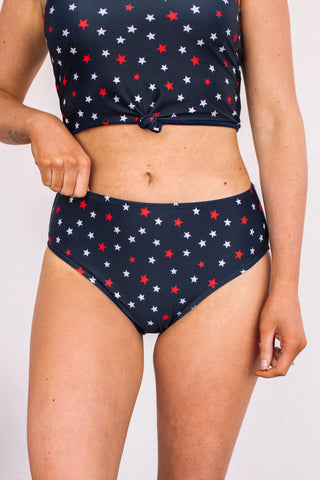 An individual is seen in a navy two-piece swimsuit adorned with red, white, and blue star patterns. The top is sleeveless with a front knot design, and they are paired with the Star Spangled Bottoms from Western Edge Boutique, which are high-waisted and crafted from a comfortable polyester spandex blend. The image highlights the torso and upper leg region.