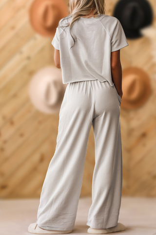 A person with long blonde hair stands with their back to the camera, dressed in a casual light gray Berkley Women Raw Hem Shirt and Wide Leg Pants Loose Set by Aapparella. The background features a wooden wall adorned with several hats, creating an inviting daily scene.