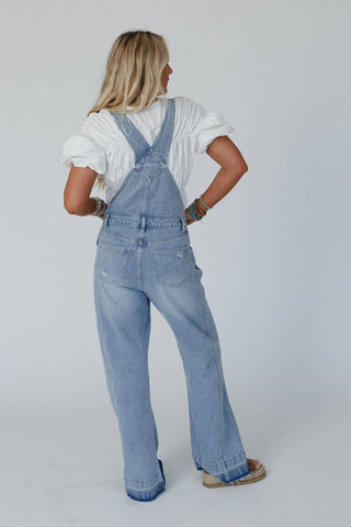 A person with long blonde hair stands facing away, wearing the Cali Dream Denim Overalls in Light Wash by Three Bird Nest, paired with a white shirt featuring puffed sleeves. Their hands rest on their hips, adorned with multiple colorful boho bracelets. The background is plain and light-colored.