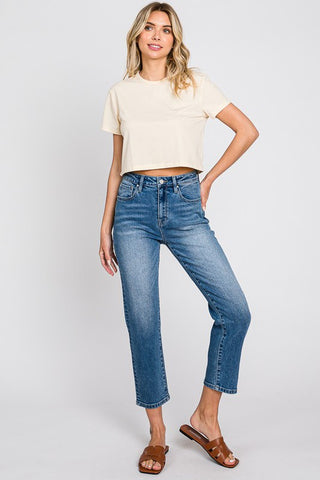 A woman stands against a plain background, wearing a light-colored, short-sleeve cropped top and the trendy Mavee High Rise Stretch Slim Crop Jeans by Petra153. She pairs her outfit with brown strappy sandals. Her right hand is placed on her hip, and she has a relaxed expression.