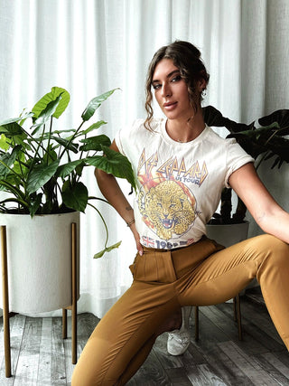 A person with wavy hair poses indoors in front of a light-colored curtain and tall potted plants. They are dressed in a Pyromania Def Leppard '83 Vintage Tee from Life Clothing Co and mustard-colored pants. They hold a confident expression, one arm raised, and the other resting on their bent knee.