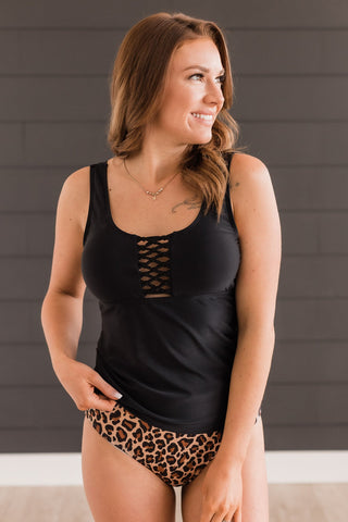 A woman with light brown hair, wearing the MACK & MAI Eastyn Tankini Top Braided Front Swimsuit Top in Black and leopard print bikini bottoms, is standing against a dark gray, horizontally-paneled wall. She is smiling and looking to her left.