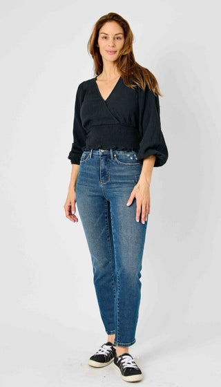 A woman stands against a plain white background wearing a black V-neck long-sleeve top and Judy Blue's Britt Slim Leg Crop Non Distressed Tummy Control Panel jeans. She has long, wavy brown hair and is smiling slightly. She is also wearing black and white sneakers and has one hand resting on her hip.