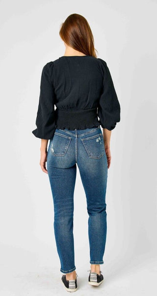 A person with long hair wearing a black long-sleeved blouse, blue jeans from the Britt Slim Leg Crop Non Distressed Tummy Control Panel line by Judy Blue, and black shoes stands with their back facing the camera against a plain white background.