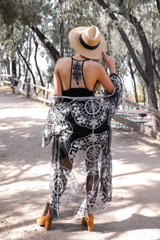 A woman wearing a straw hat, black strappy top, and the Riley Ruffled Trim Tattoo Mesh Back Slip Dress by Leto Collection, featuring a long sheer black lace cover-up with floral patterns, walks away on a sunlit path lined with trees and a wooden fence. Embracing her bold style, she is holding one hand to her hat and wearing brown ankle boots.