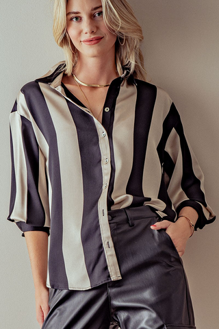 A woman stands confidently against a neutral background, wearing the Linden Bold Stripe Drop Shoulder Button Down Shirt from Trend Notes. The stylish black and beige vertically striped blouse, with the top button undone, is paired with black leather pants. She has blonde hair and accessorizes with a gold necklace and bracelet, one hand casually tucked into her pocket. The daring stripes enhance her chic look.