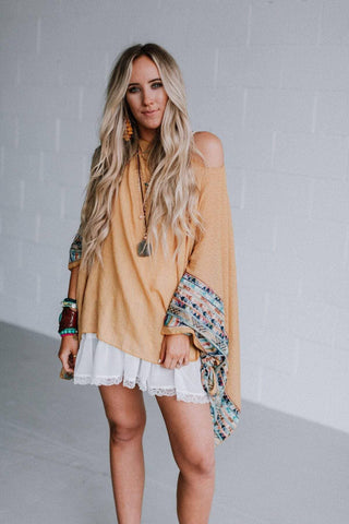 A woman with long blonde hair is standing against a plain white wall. She is wearing the Fallon Embroidered Sleeve Poncho in mustard from Western Edge Boutique, paired with white lace-trimmed shorts, multiple bracelets, and necklaces. One shoulder of her poncho is exposed, giving a casual look.