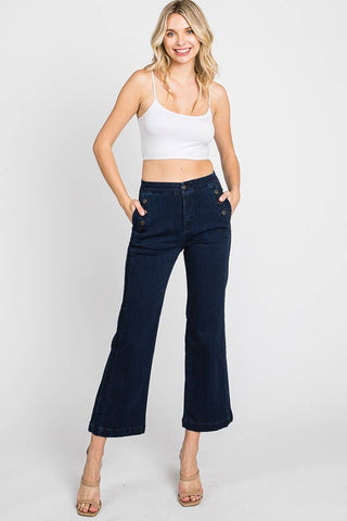 A woman with blonde hair is smiling and standing against a plain white background. She is wearing a white crop top, dark blue Petra153 Pistol Wide Leg Sailor Jeans, and nude-colored heels. Her hands are in her pockets, and she stands confidently.