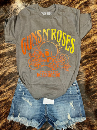 A stylish vintage look is effortlessly created with the Tilley Rocker Queen Tee by Western Edge Boutique, which features a light gray color and a striking yellow and orange "Guns N' Roses" logo above a skull adorned with roses. Pair it with distressed blue denim shorts, all laid out on a brown, textured surface.