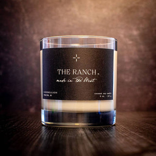 A glass candle with a black label sits on a dark wooden surface. The label reads "THE RANCH, made in the West" and "COCONUT SOY CANDLE." Made by R. Rebellion in Houston, TX, it weighs 8 oz (227 g). This all-natural wax Sandalwood candle is housed in a reusable whiskey glass.