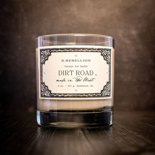 A hand-poured candle in a clear glass container sits on a dark surface. The label reads "R. Rebellion" at the top, followed by "Coconut Soy Candle," "Dirt Road Candle 8 oz.," and "Made in the West" with "227 g" at the bottom, bordered by an ornate design, showcasing its all-natural wax.