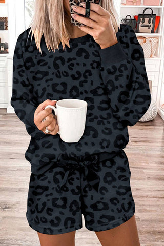 A person with blonde hair stands indoors, holding a white mug in one hand and taking a mirror selfie with the other. They are wearing a matching dark leopard print sweatshirt and shorts set from the stylish *Lovesoft* Wells 2 Piece Leopard Loungeseat collection. The background features white shelves and drawers.