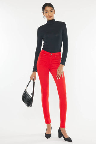 A person with their hair tied back is posing against a white background, wearing a black turtleneck and Smoke Show Red Skinny Jeans by Kan Can USA. They hold a black handbag and wear black heels.