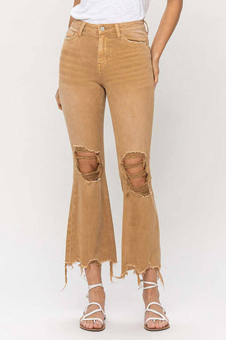 A person is wearing VERVET by Flying Monkey's Britton Vintage High Rise Crop Flare Distressed jeans in light brown, featuring a frayed hem and distressed rips at the knees. They have paired the jeans with white strappy sandals and a tucked-in white top, standing against a neutral background.