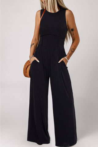 A person with long blonde hair is wearing the Atley Black Sleeveless High Waist Wide Leg Jumpsuit from Little Daisy Closet and has their hands in the pockets. They have tattoos on their right arm and are holding a round woven handbag on their left arm. The versatile styling options of this outfit make it suitable for various occasions. The background is plain and light-colored.