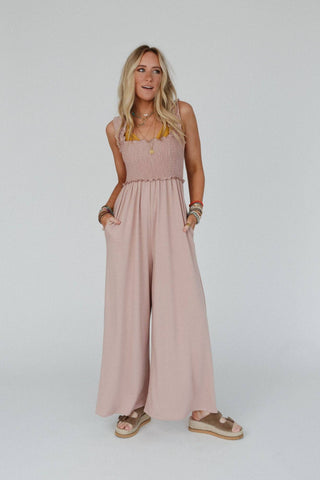 A woman stands against a plain background, wearing the Colt Smocked Ruffle Jumpsuit in blush-pink from Three Bird Nest. The sleeveless jumpsuit features flowy pant legs and smocked ruffle details. She has long blonde hair and accessorizes with multiple bracelets and layered necklaces, completing her look with comfortable wedge sandals.