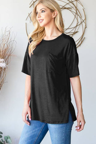 A woman with long blonde hair is standing indoors wearing the QUINNIE ROUND NECK SOLID TOP by Western Edge Boutique, a black, half-sleeve top with a pocket on the left side, and blue jeans. She is looking to her right and smiling. The background features decorative wooden sticks and circular wall art.