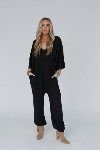 A woman with long blonde hair stands against a plain backdrop, wearing the Lovely As Ever Harem Jumpsuit in charcoal from Three Bird Nest, her hands tucked into its loose-fitting pockets. She accessorizes with multiple boho bracelets on both wrists and finishes the look with comfy beige ankle boots.