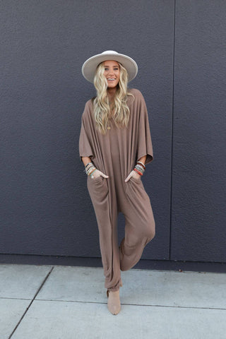 A woman stands against a dark gray wall, smiling, with her hands in the pockets of her Lovely As Ever Harem Jumpsuit by Three Bird Nest. She is wearing a loose-fitting light brown outfit made from comfortable knit fabric, a wide-brimmed hat, and several bracelets. Her long, wavy blonde hair cascades over her shoulders as she stands on a paved walkway.