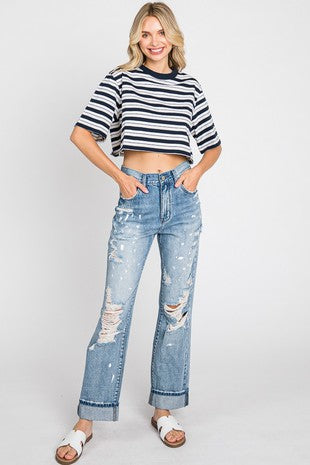 A woman stands against a plain backdrop, smiling. She is wearing a black and white striped short-sleeve cropped top and high-waisted Dolly Distressed Paint Splatter Jeans from Petra153, which feature rolled-up cuffs. Made of 100% cotton, her jeans have a distressed hem. She has long wavy blonde hair and is wearing white slide sandals.