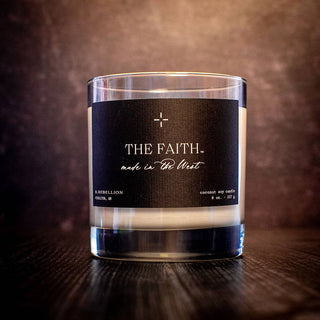 A glass jar candle labeled "The Faith Candle 8 oz.," made in the West, featuring a sleek black label with white text and decorative elements. Described as "R. Rebellion Portland, OR" and "coconut soy wax candle 8 oz | 227 g," it also boasts a wood wick. The background is dark with a textured effect.