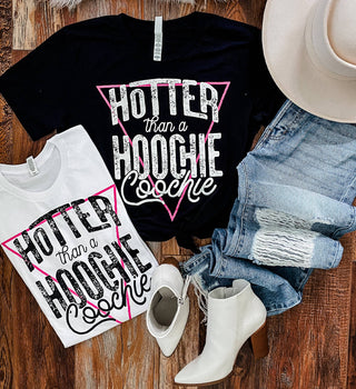 Two outfits are laid out on a wooden floor, perfect for a country concert. One features the Western Edge Boutique's "Jackson Hotter than a Hoochie Black Tee," paired with denim shorts, white boots, and a white hat. The other includes a white T-shirt with the same text, blue ripped jeans, and white boots.