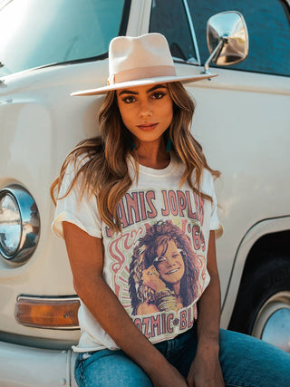 A woman with long, wavy hair wearing a light-colored fedora hat and a vintage Janis Joplin Kozmic Blues Vintage Tee from Western Edge Boutique leans against a white retro van. The relaxed crew neck tee showcases a colorful graphic of Janis Joplin. The background is a clear, sunny day.