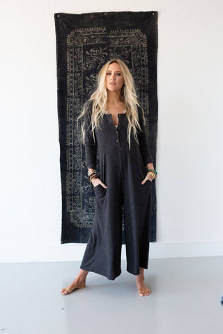 A woman with long, blonde hair stands barefoot indoors on a light floor, confidently wearing the Simply Comfort Jumpsuit in dark gray by Three Bird Nest. She has her hands in her pockets and is positioned in front of a large, intricate dark-colored tapestry hanging on the wall behind her.
